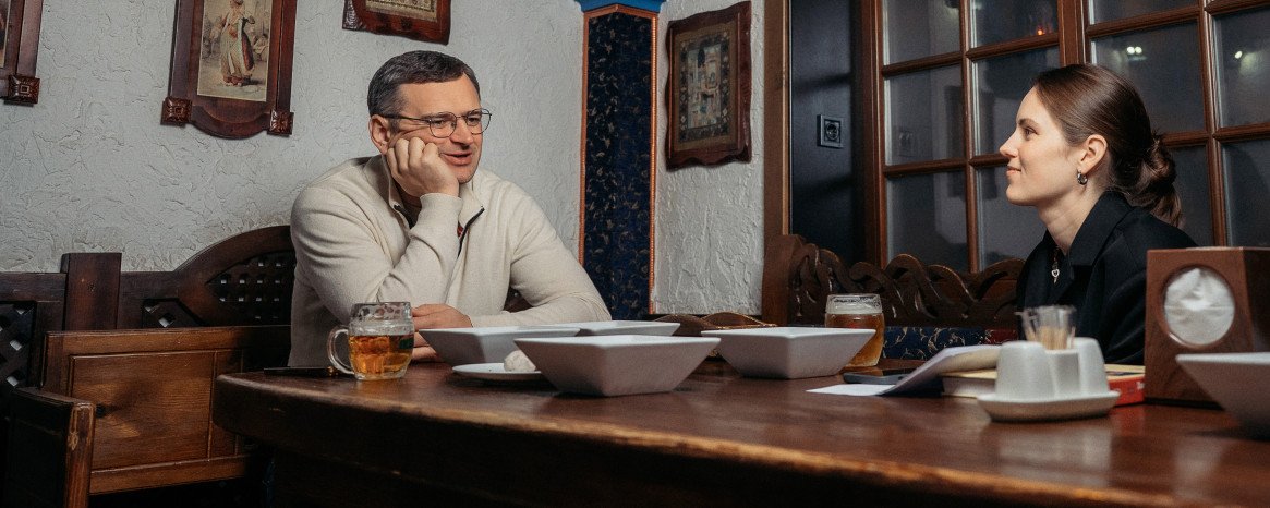 Журналістка сидить за столом з Дмитром Кулебою у луцькому закладі "Кораблик" у залі, стилізованому як каюта корабля, на столі білі тарілки зі стравами.