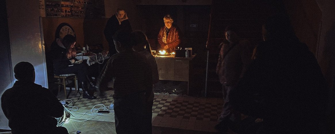 Темне приміщення, у центрі жінка, підсвічена свічками, навколо темні силуети людей.