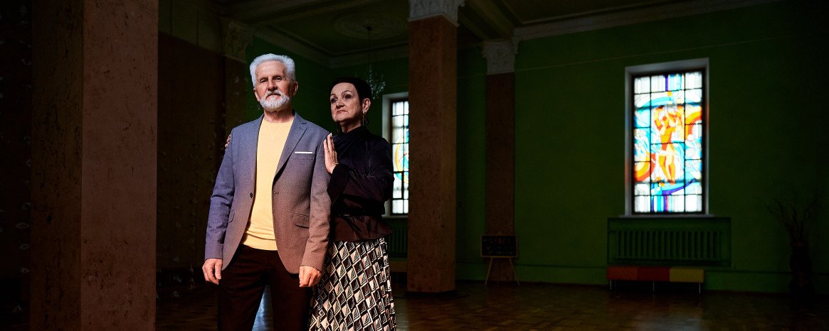 Сивий чоловік з бородою у костюмі та стильна жінка з короткою стрижкою ніжно сперлись один на одного у темному залі з вітражними вікнами.