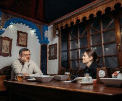 Журналістка сидить за столом з Дмитром Кулебою у луцькому закладі "Кораблик" у залі, стилізованому як каюта корабля, на столі білі тарілки зі стравами.