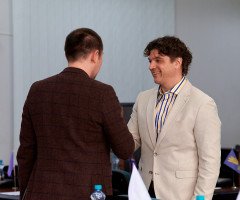 Двоє чоловіків тиснуть один одному руки. Один у темно коричневому піджаку, інший у молочному.