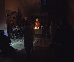 Темне приміщення, у центрі жінка, підсвічена свічками, навколо темні силуети людей.