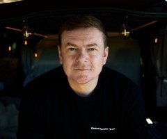 Портрет Святослава Савича. По груди. Він у чорній футболці, сидить всередині будинку на колесах, позаду горять вогники.