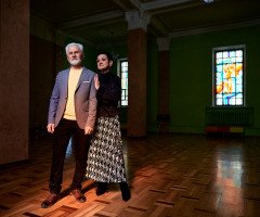 Сивий чоловік з бородою у костюмі та стильна жінка з короткою стрижкою ніжно сперлись один на одного у темному залі з вітражними вікнами.