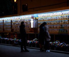 Люди стоять перед фотостендом із портретами загиблих у війні з Росією. На вулиці темно, стенд підсвічується. Луцьк. Театральний майдан.