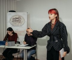 Дівчина з червоним пасмом волосся говорить жестикулюючи на тлі двох слухачів.