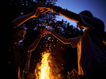 Двоє людей тримаються за руки на фоні ватри та синього вечірнього неба.