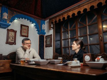 Журналістка сидить за столом з Дмитром Кулебою у луцькому закладі "Кораблик" у залі, стилізованому як каюта корабля, на столі білі тарілки зі стравами.