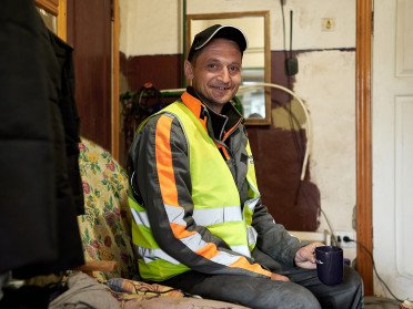 Чоловік у спецодязі та кепці сидить у невеликій сторожовій комірчині.