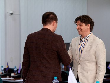 Двоє чоловіків тиснуть один одному руки. Один у темно коричневому піджаку, інший у молочному.