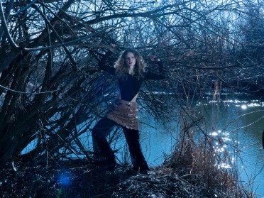 Кучерява дівчина стоїть на березі водойми, підтримує руками гілки дерева