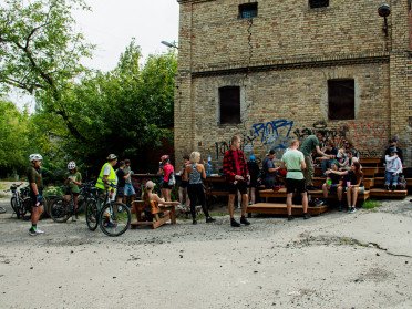 Група велосипедистів на тлі промислової будівлі.
