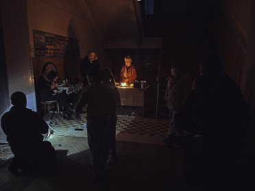 Темне приміщення, у центрі жінка, підсвічена свічками, навколо темні силуети людей.