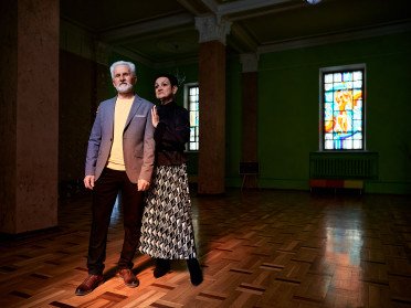 Сивий чоловік з бородою у костюмі та стильна жінка з короткою стрижкою ніжно сперлись один на одного у темному залі з вітражними вікнами.