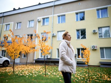 Людмила Головенко. Струнка жінка в білій куртці та чорних штанах на тлі лікарні та пожовтілих дерев.