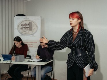 Дівчина з червоним пасмом волосся говорить жестикулюючи на тлі двох слухачів.