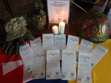 Стіл, на якому лежить військова каска, піксельний капелюх, рукавиці, записки з іменами полоеглих військових та горить дві свічки.