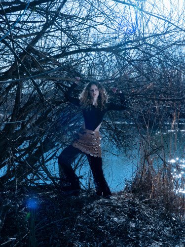 Кучерява дівчина стоїть на березі водойми, підтримує руками гілки дерева