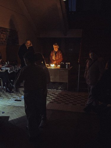 Темне приміщення, у центрі жінка, підсвічена свічками, навколо темні силуети людей.