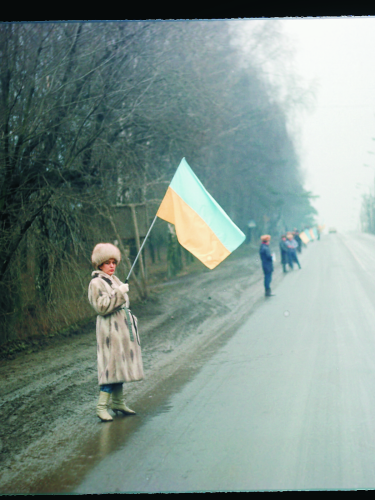 Жінка на узбіччі дороги стоїть з синьо-жовтим прапором, позаду неї люди вздовж дороги, на дорозі одиноке авто.