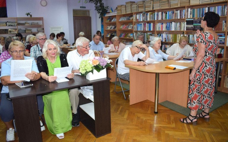 Люди сидять за столами в приміщенні бібліотеки, слухають лекторку
