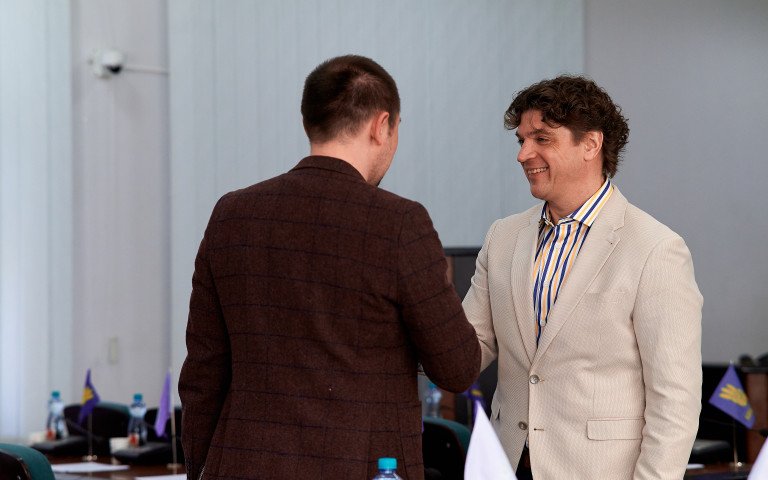 Двоє чоловіків тиснуть один одному руки. Один у темно коричневому піджаку, інший у молочному.