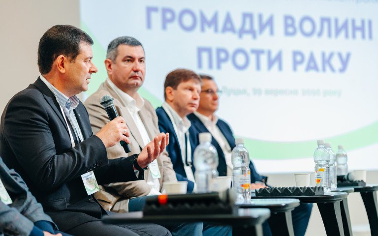 Чоловіки з мікрофонами на тлі банеру «Громади - Волині проти раку».