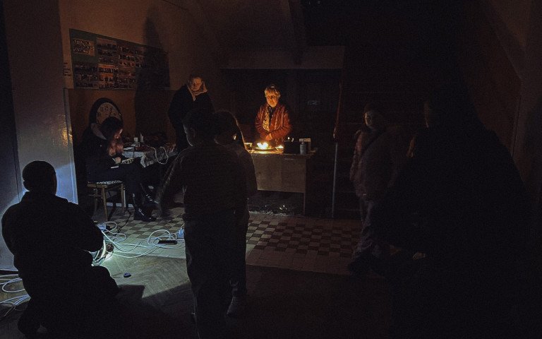 Темне приміщення, у центрі жінка, підсвічена свічками, навколо темні силуети людей.