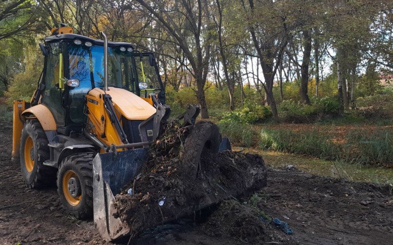 трактор згрібає рослинні залишки і сміття на території зеленої зони біля водного каналу