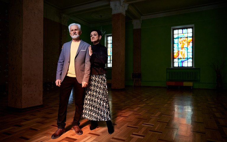 Сивий чоловік з бородою у костюмі та стильна жінка з короткою стрижкою ніжно сперлись один на одного у темному залі з вітражними вікнами.