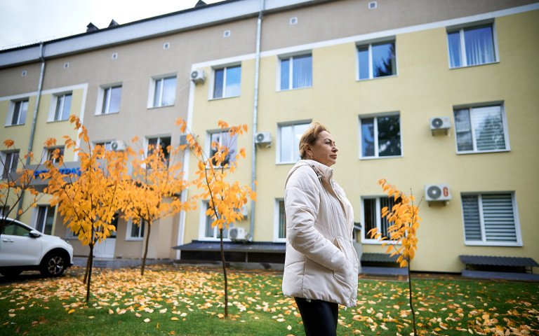 Людмила Головенко. Струнка жінка в білій куртці та чорних штанах на тлі лікарні та пожовтілих дерев.