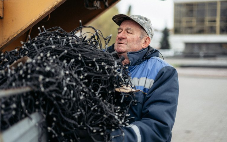 Микола Іванович розплутує десятки метрів ялинокових гірлянд, аби створити місту свято. Літній чоловік у спецодязі несе оберемок гірлянд.