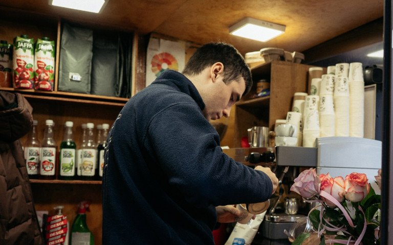 Всередині невеликого кава-кіоску хлопець біля кавомашини готує капучино.