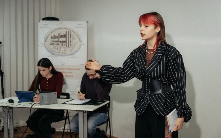 Дівчина з червоним пасмом волосся говорить жестикулюючи на тлі двох слухачів.
