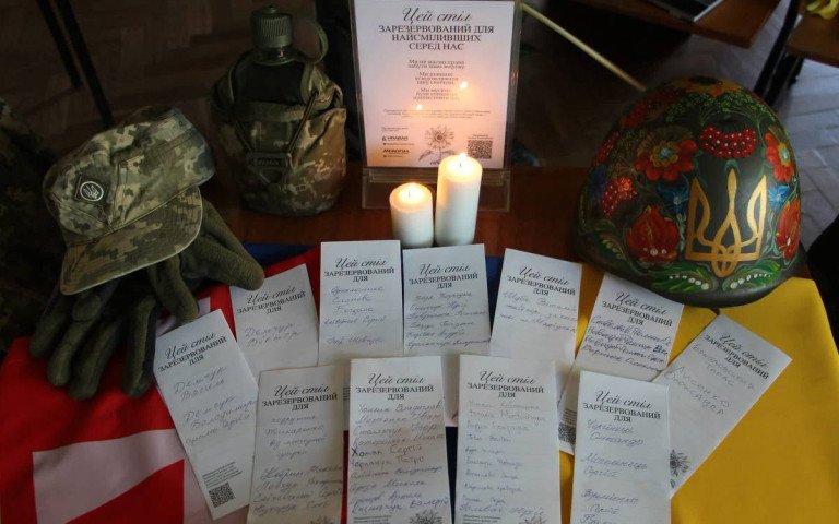 Стіл, на якому лежить військова каска, піксельний капелюх, рукавиці, записки з іменами полоеглих військових та горить дві свічки.